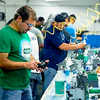 Several workers in a factory setting assemble parts at a workstation; one man in the foreground uses a handheld device while wearing ear protection.