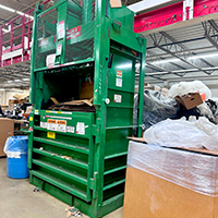 A green industrial cardboard baler in a warehouse surrounded by boxes, plastic wrap, and a blue trash bin.